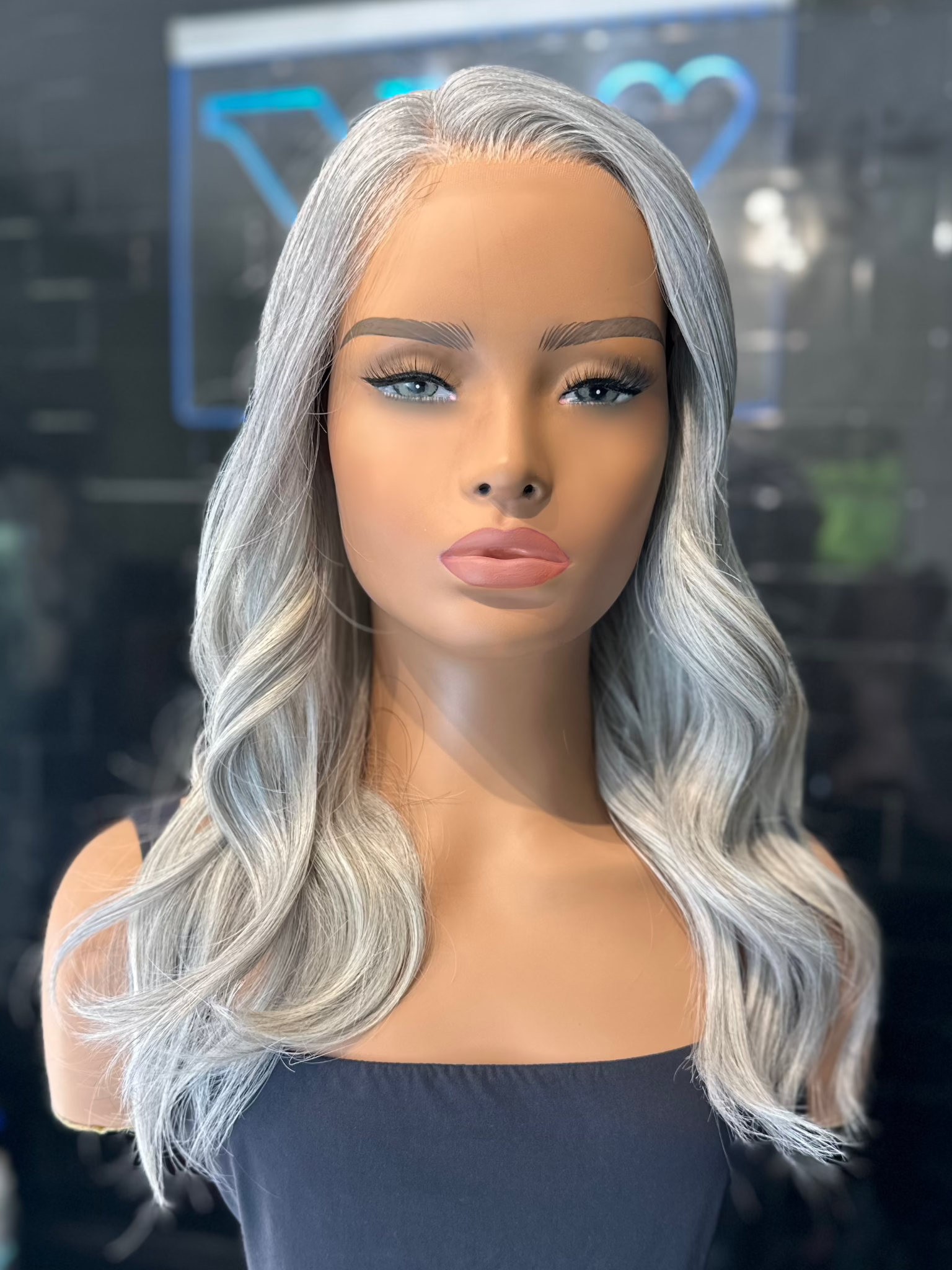 A mannequin head displaying a light grey, salt and pepper wig with a white base and charcoal peppered throughout. The wig has a textured cut that gives a lightweight feel and is styled with a silicone grip.